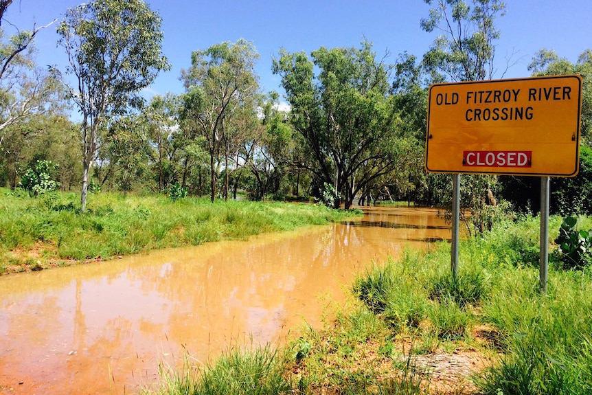 Brown water covers the flooded Old Fitzroy River crossing with a yellow sign on the right.