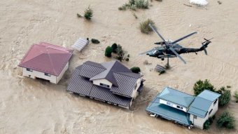 An army helicopter flies above a group of three houses, surrounded by rushing flood waters.