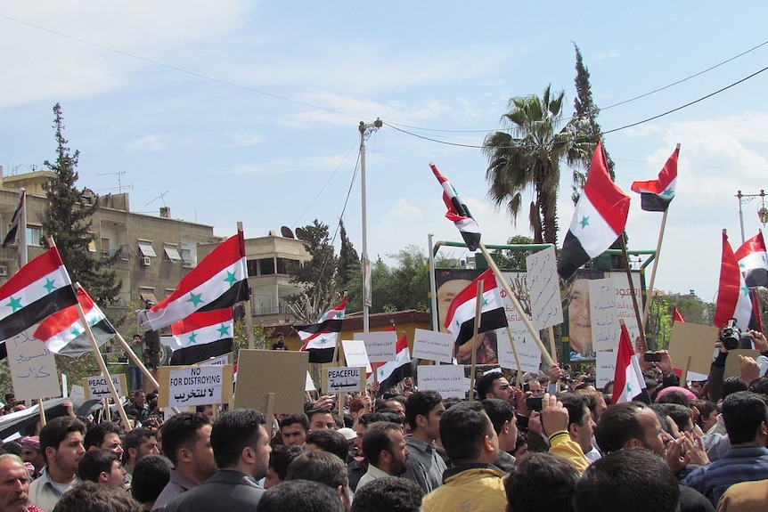 syrian flags are waved by a crowd of young men in a town square holding anto-war placards