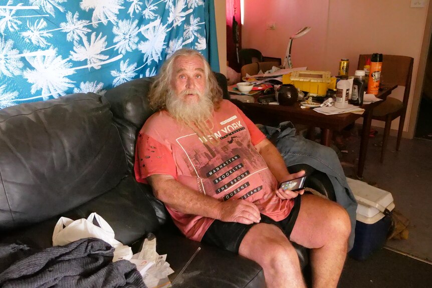 A man with long white hair and a beard, sitting on a black couch.