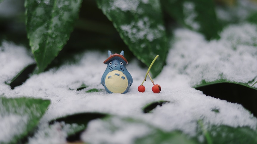 A figurine of the animated character Totoro sits on a leaf in the snow. Two berries sit next to him.