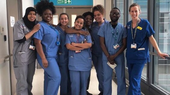 A group of people in various shades of blue and grey scrubs, standing in a hospital hall.
