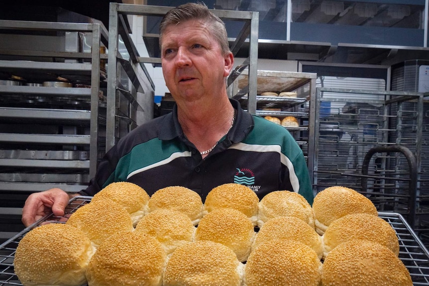 A man holding a tray of bread rolls with metal bakery racks behind him.