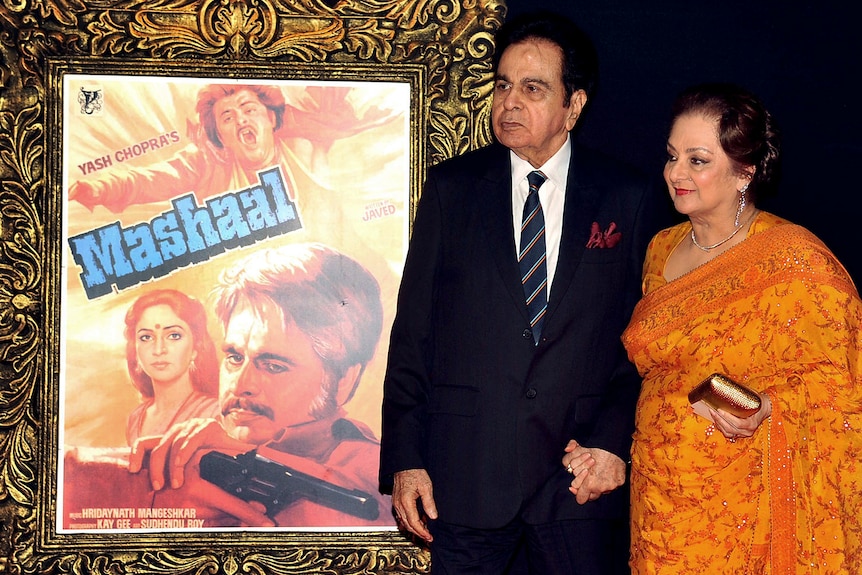 Dilip Kumar and his wife Saira Banu pose in front of a movie poster on the red carpet.