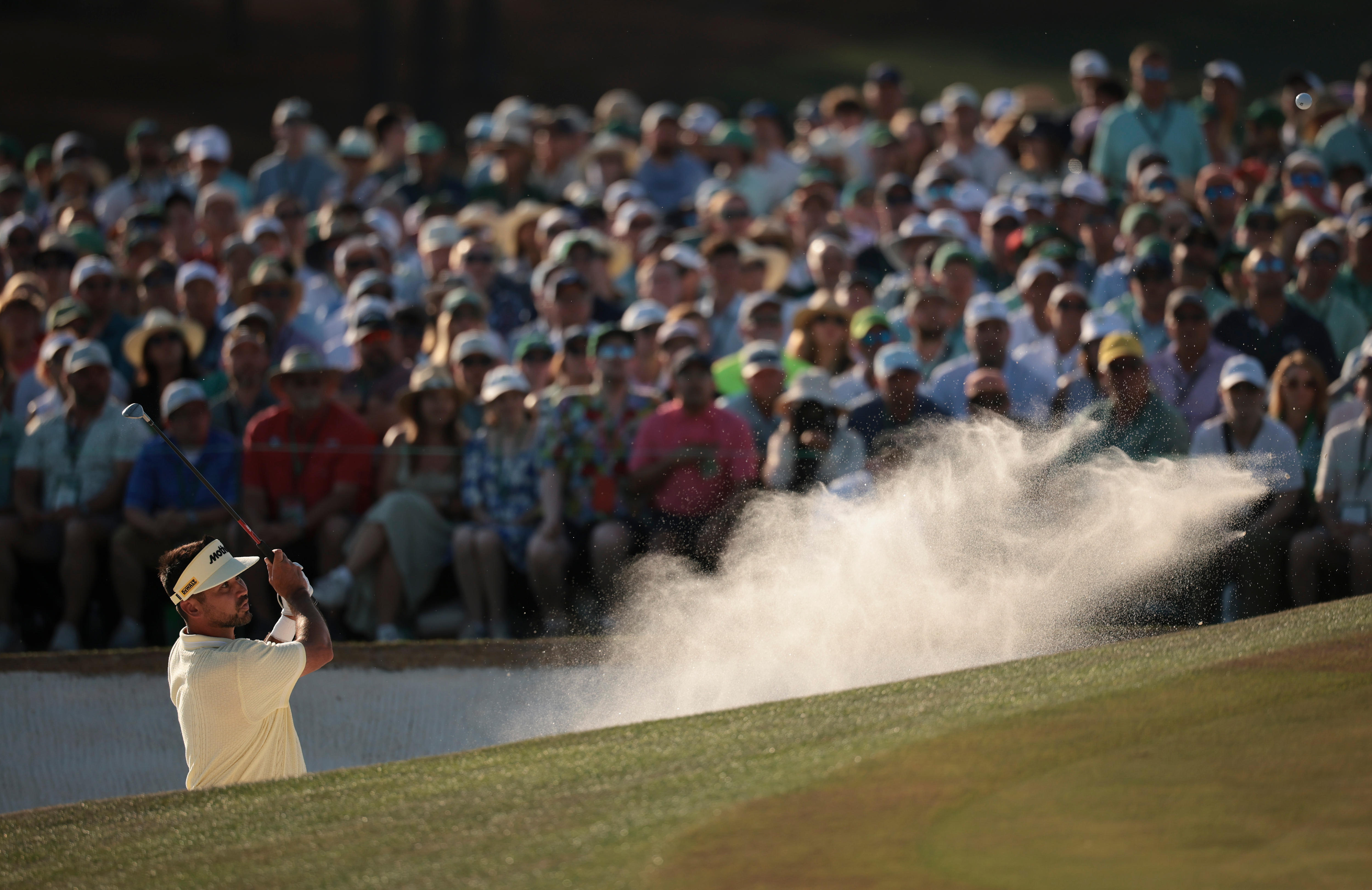 A golfer hits a ball out of a bunker, with a wave of sand captured flowing through the air from the shot.