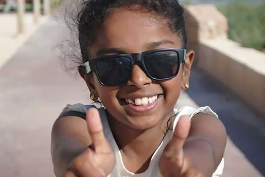 Une tête et des épaules d'une jeune fille souriante, portant des lunettes de soleil et tenant deux pouces vers le haut