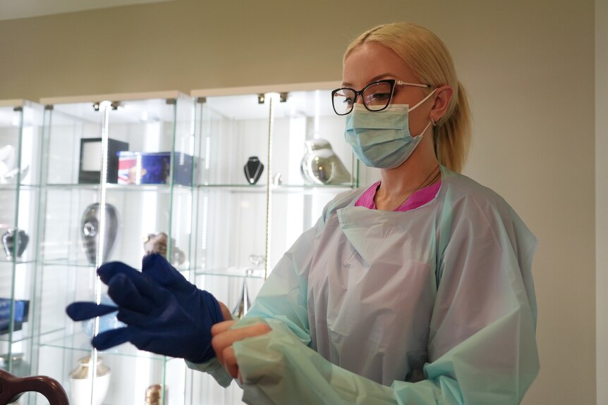 A young woman wearing pink scrubs and a blue protective gown puts on gloves.