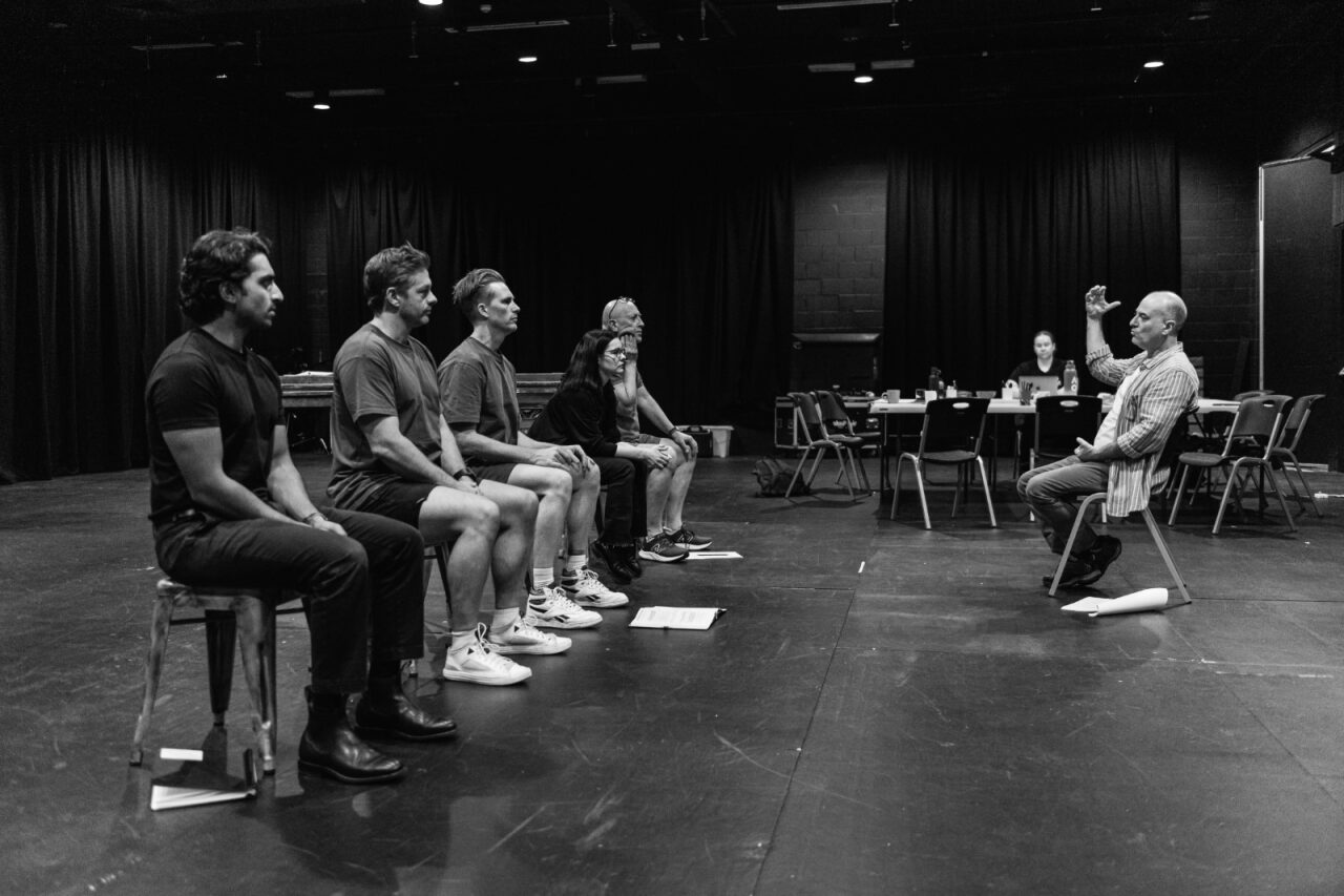 Cast rehearsal. One person sits on a chair facing a row of people.