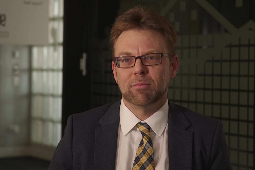 Head shot of Dr Paul Blacklow sitting for an interview, wearing grey suit and yellow and blue tie