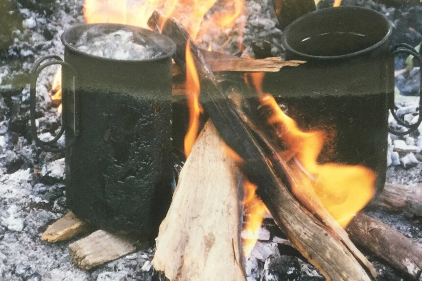 Two small black metal 'quart pots' sit next to a small fire with water in them.  The water in one is boiling.