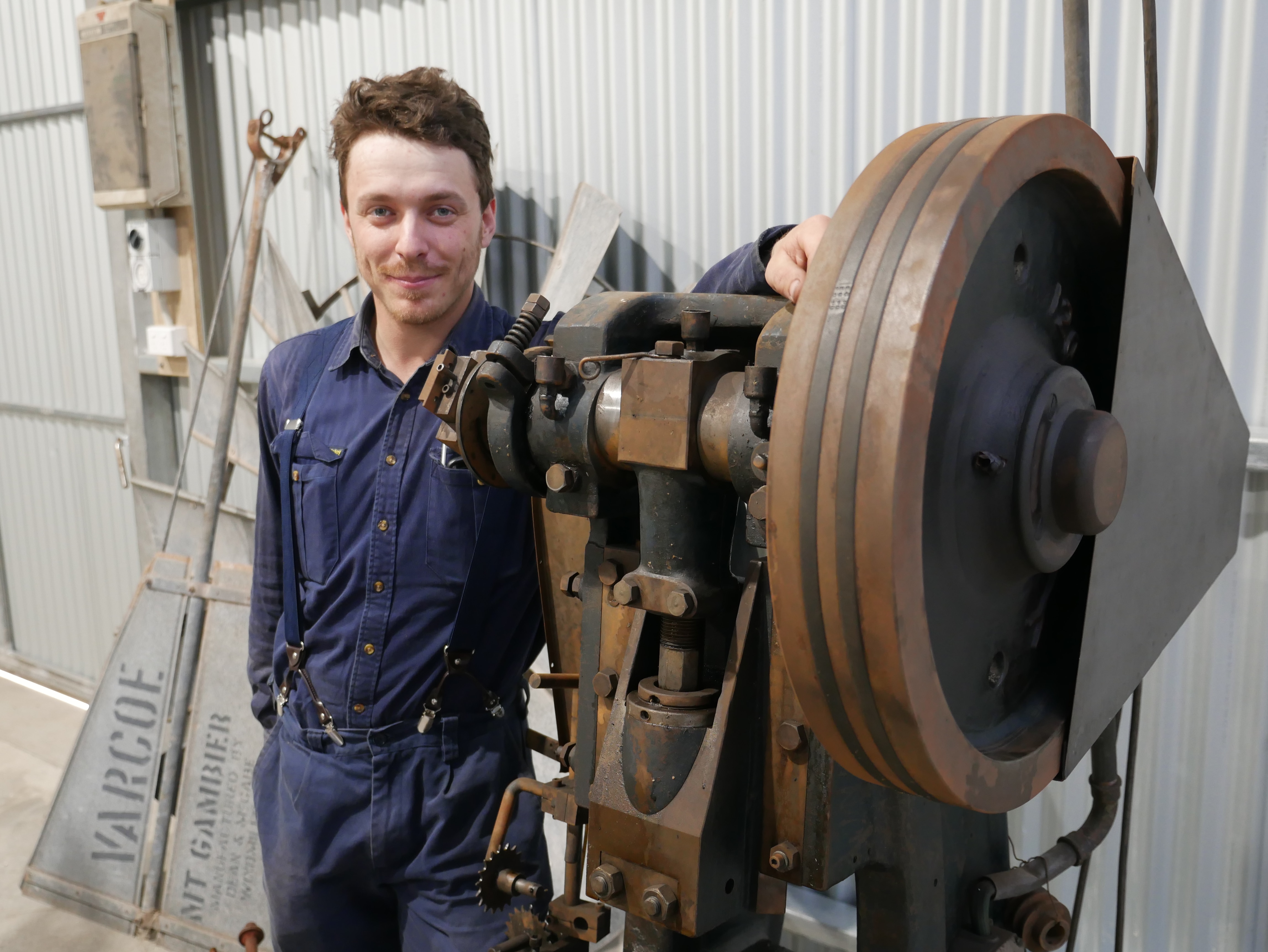 A man wearing blue mechanic's clothes standing next to a large machine. 