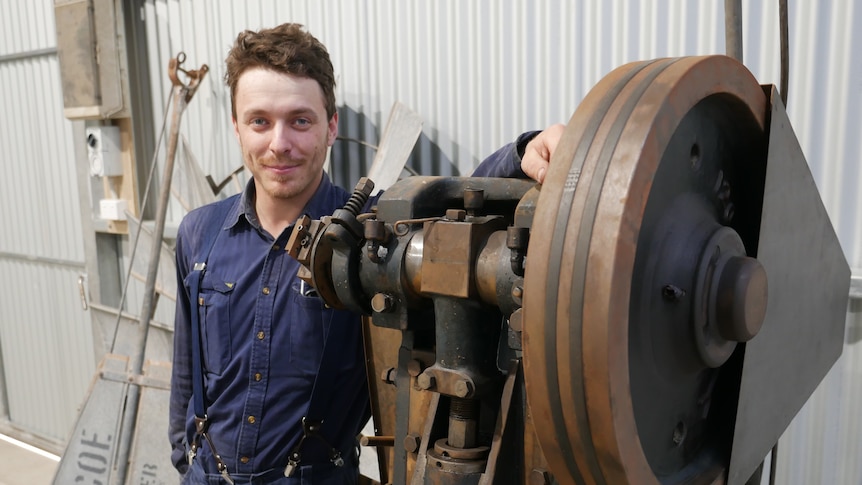 A man wearing blue mechanic's clothes standing next to a large machine. 