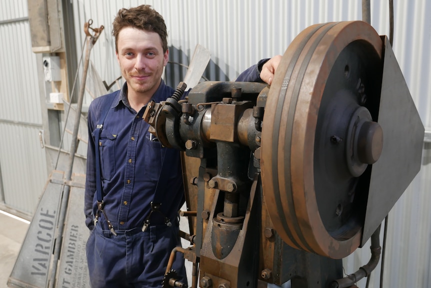 A man wearing blue mechanic's clothes standing next to a large machine. 