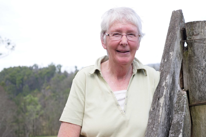 A woman in her 70s standing by a wooden fence post, white sky and distant trees behind her.