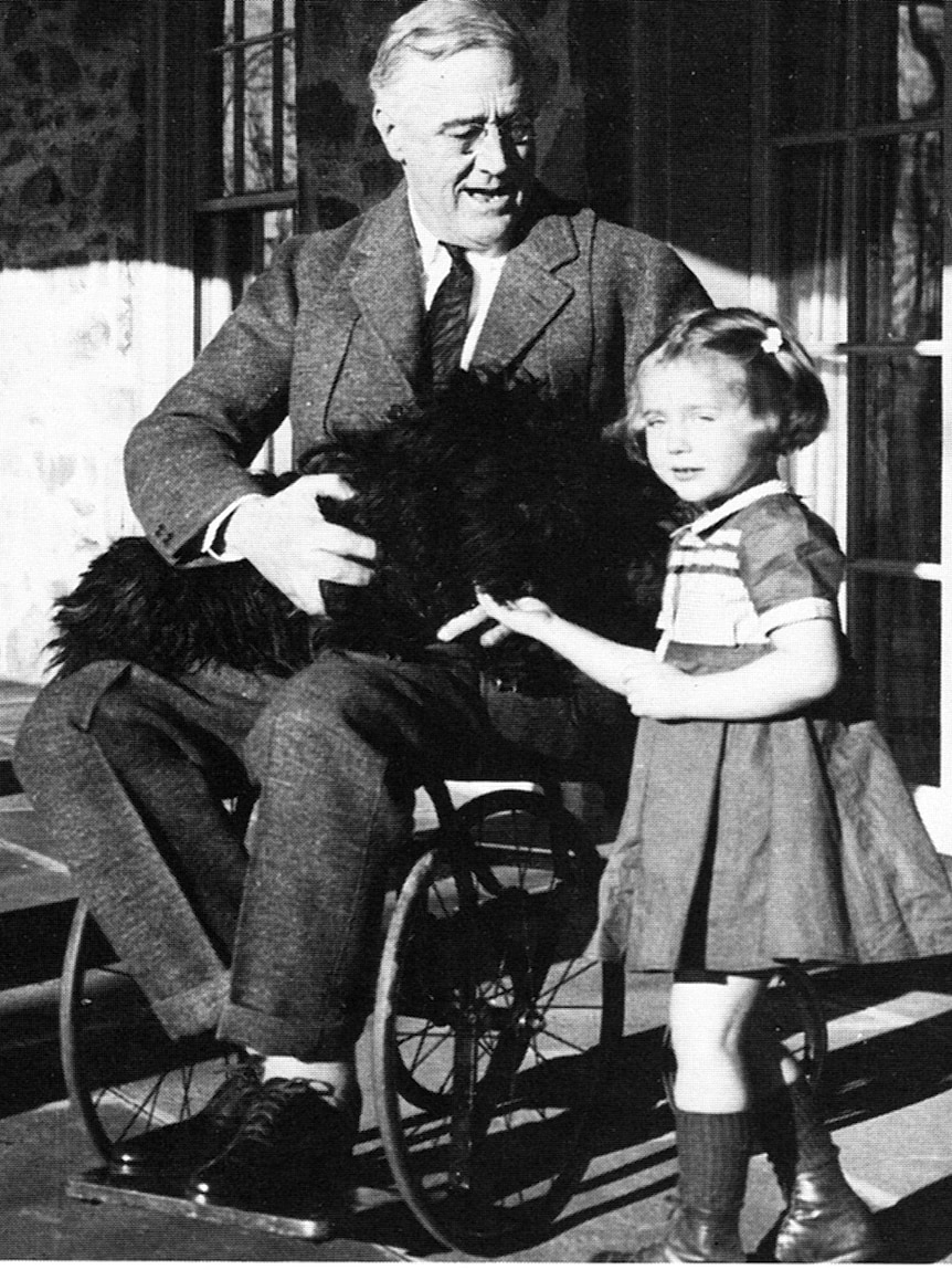 Historic black and white image of President Roosevelt sitting on wheelchair with dog on lap and little girl standing