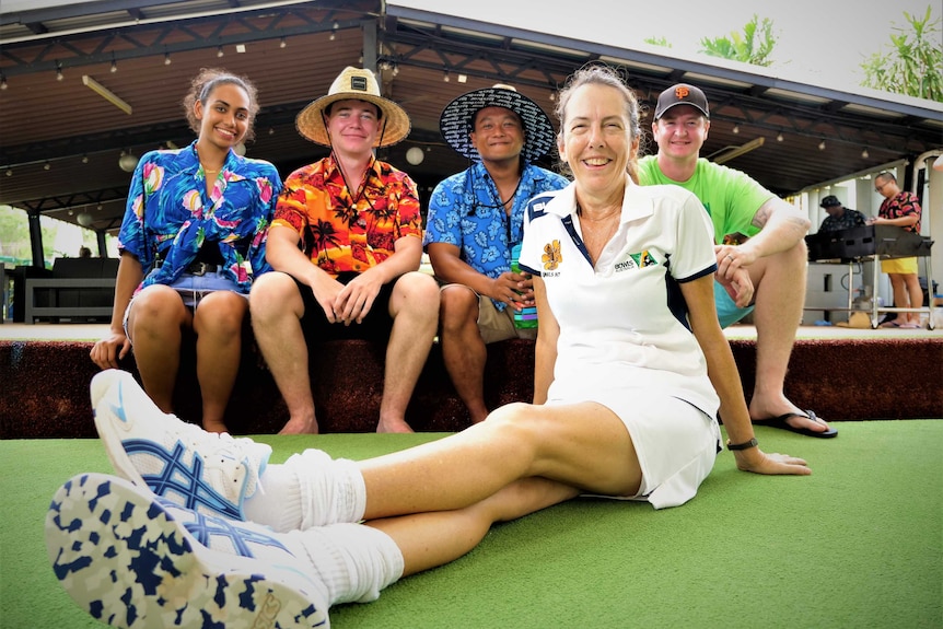 Woman sitting on artificial bowling green with long socks pulled down. Three boys and a girl behind in loud shirts.