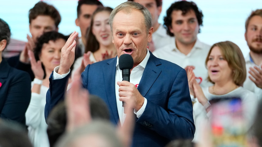 Donald Tusk speaking into a microphone surrounded by a crowd clapping. 