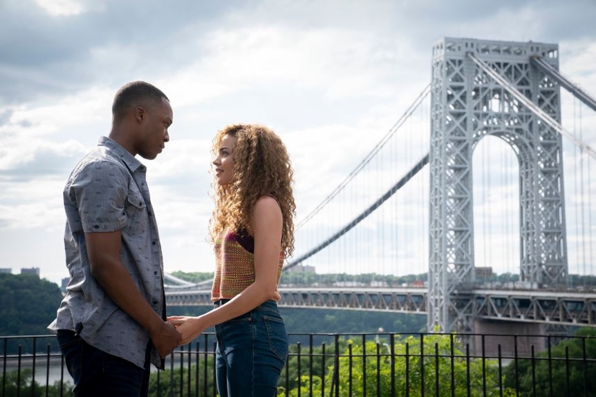 A man and a woman stand hand in hand with a large bridge behind them.
