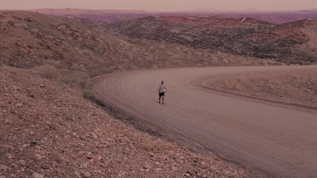 Wide shot of Matt Napier walking along a road in the desert.