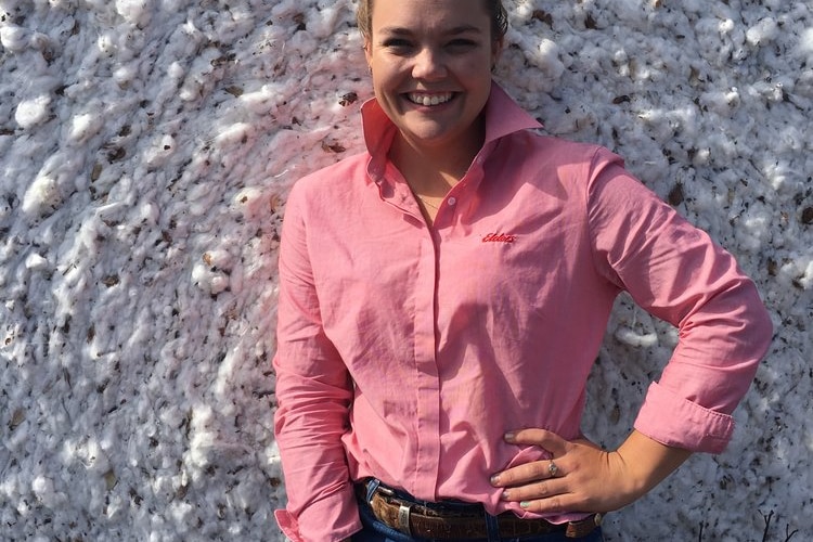 a woman in jeans and a pink log sleeve shirt standing in front of a cotton bale 