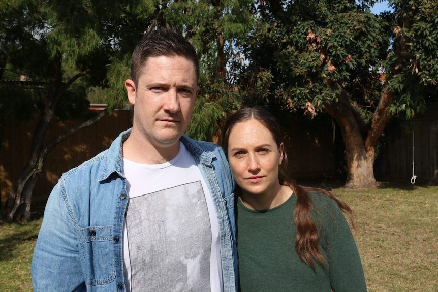 A couple stand in a park on a sunny day with serious expressions.