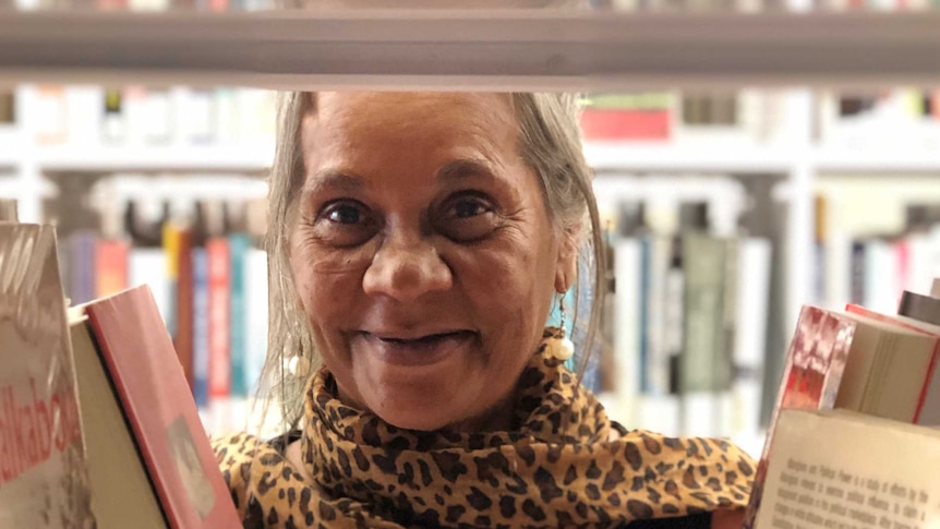 Ms Moreton smiles at camera, through a gap of books in the aisle of a library.