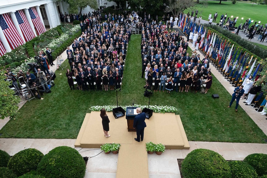 Donald Trump stands to the right of Amy Coney Barrett on a platform on grass with hundreds of people sat in front of them.