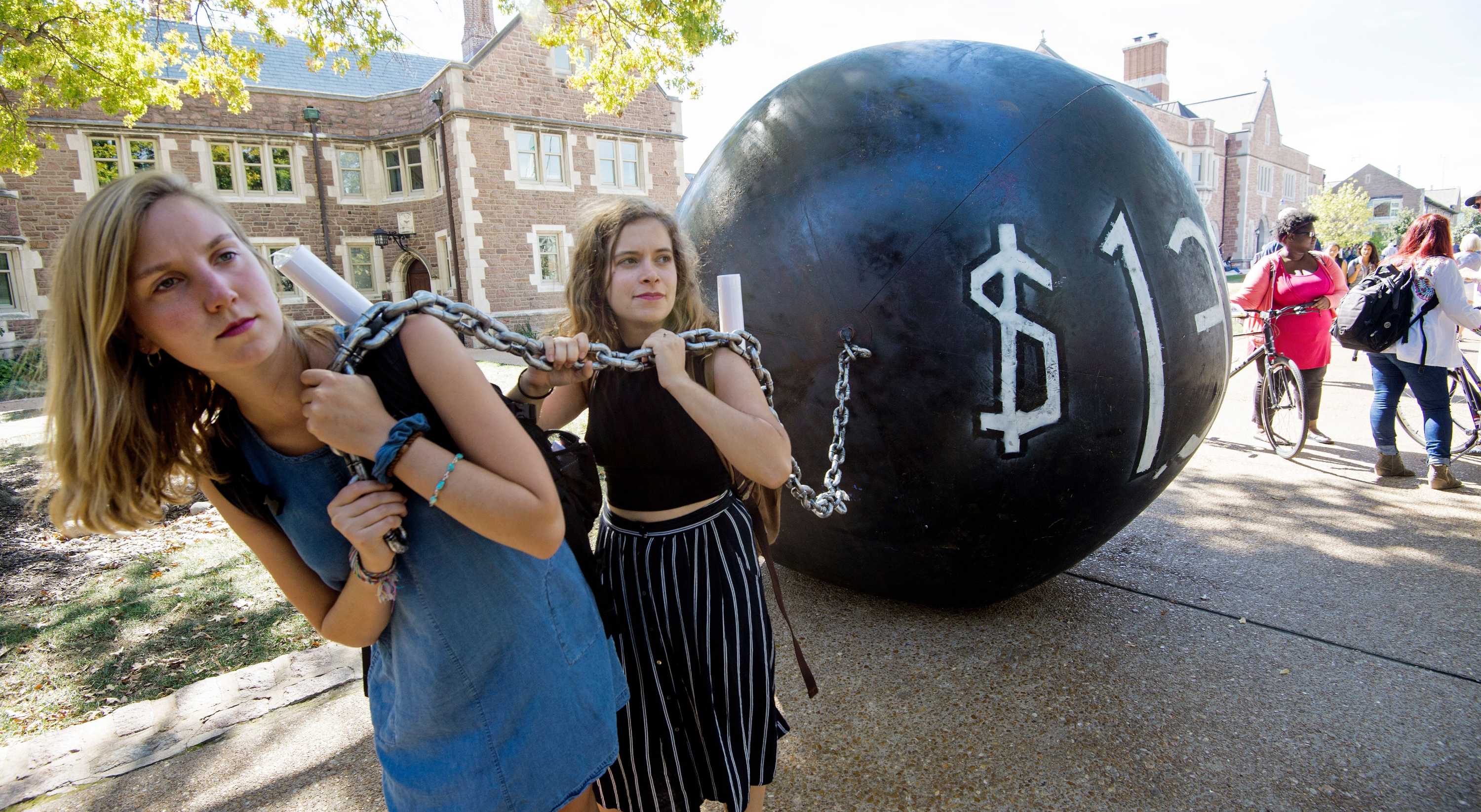 Two young women carry a large ball and chain to represent the burden of student loans