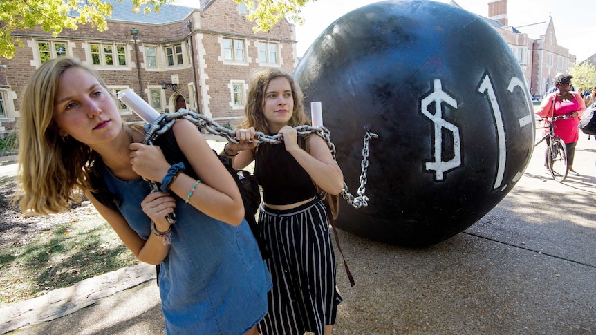 Two young women carry a large ball and chain to represent the burden of student loans