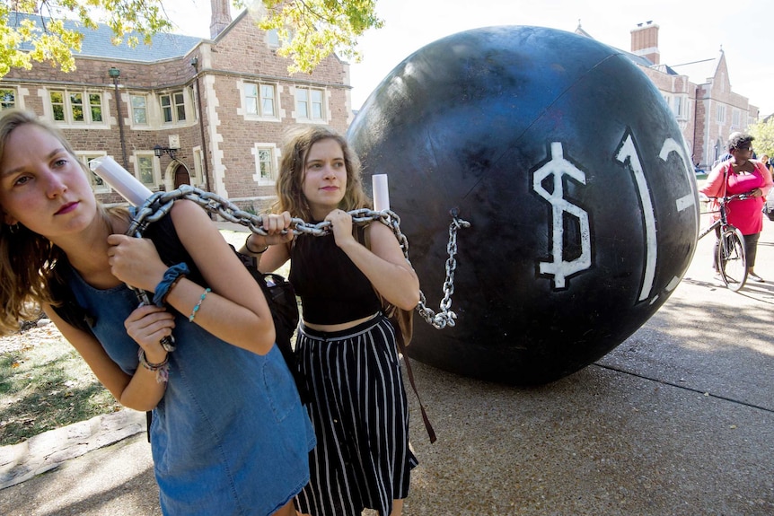 Two young women carry a large ball and chain to represent the burden of student loans