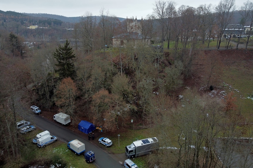 An aerial shot of forests with police vans parked along the road and a old castle building behind. 