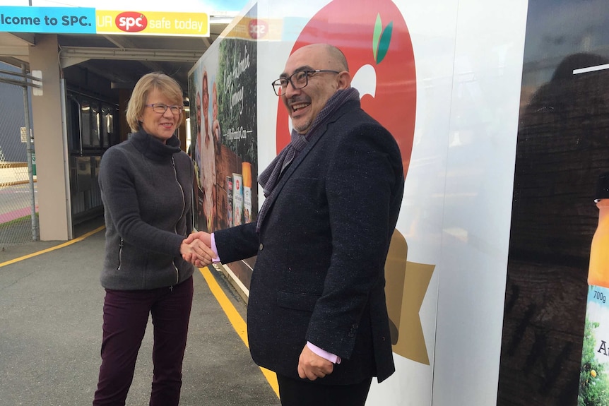 A woman with short blonde hair and a man with a shaved head smile as they shake hands in front of an SPC logo.