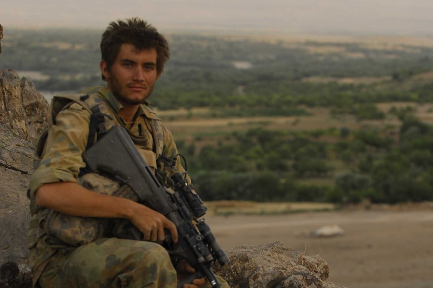 Soldier wears green camouflage and sits holding rifle amid desert landscape