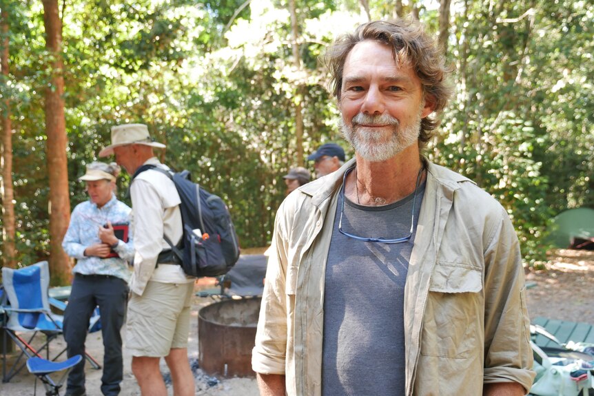 Man witj grey beard, crumpled shirt, blue tee, glasses hanging from neck smiles at camera with bushwalkers in background. 