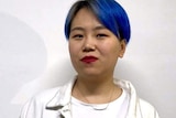 A woman with blue dyed hair wears a white jacket and stands next to a white wall