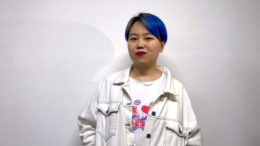 A woman with blue dyed hair wears a white jacket and stands next to a white wall
