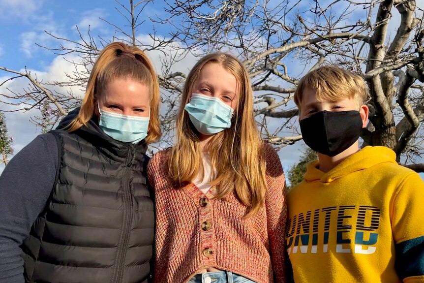 A mother and her two children, all wearing face masks and looking at the camera on a sunny day.
