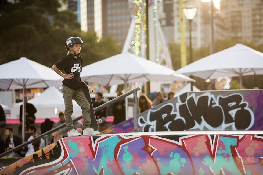 A young child in black shirt, khaki pants, helmet skateboard stands on a pink, blue, purple spraypainted skate ramp with board