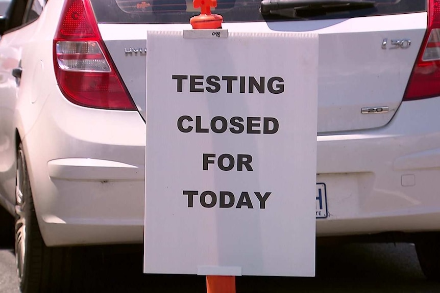 A white sign reads 'Testing closed for today', behind a queue of cars at a COVID-19 testing site.