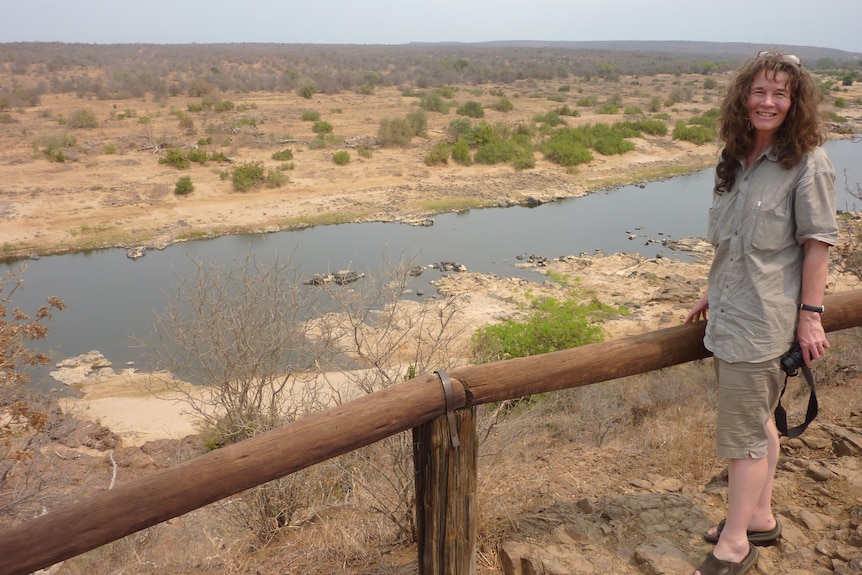 A woman, wearing khaki, standing above a water hole holding a camera 