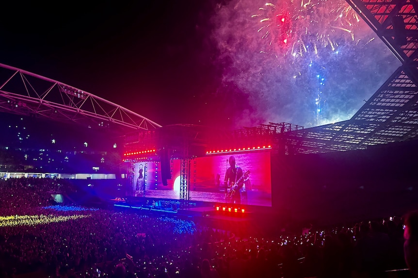 View of the stage at a rock concert from the stands, with fireworks above.