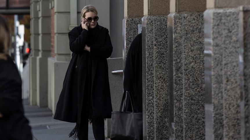 Woman in dark glasses and long black coat walks outside court while on mobile phone.