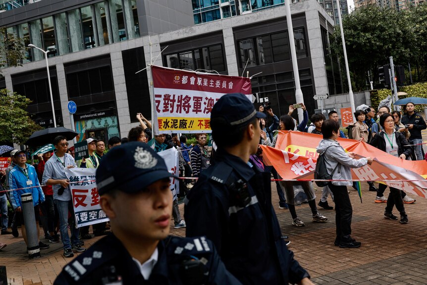 Two Asian police officers stand in front of colourful protest with people holding Chinese-language signs on city street