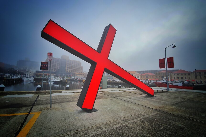 A large, red illuminated cross lies on its side with buildings, water and boats in the background.