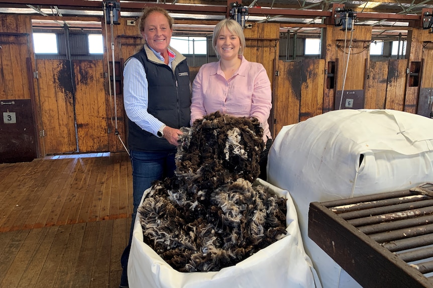 Two women stand in a shearing shed with a bale of black wool