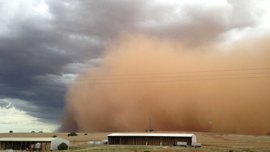 A dust storm, coloured orange, moves across a rural landscape, with farm buildings in the foreground.