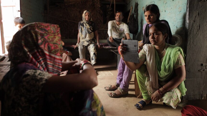 A female Indian journalist films a woman on her smart phone while her colleague and the woman's family watches