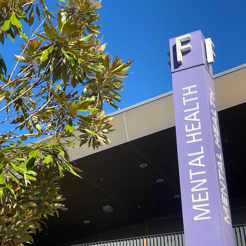 Looking skywards up a purple pillar with a sign identifying the Mental Health F block with a green leafy tree to the side