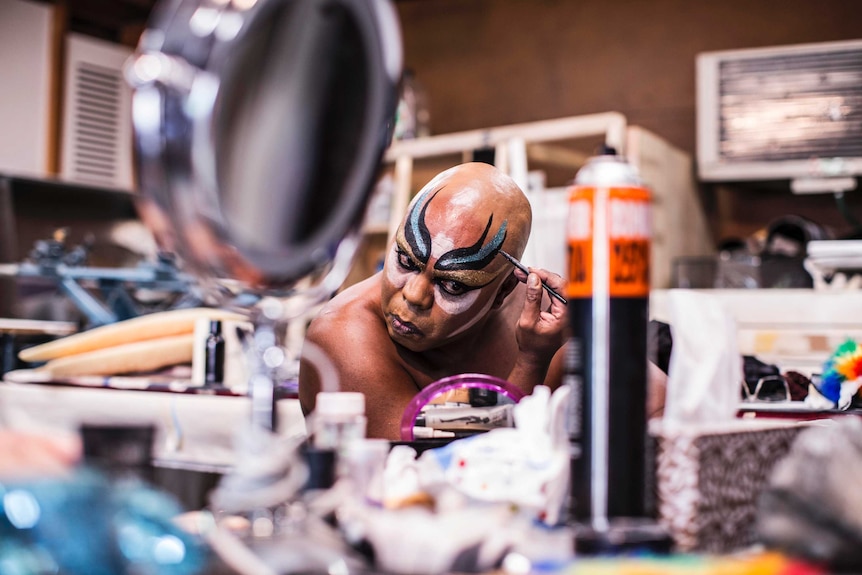 Colour photograph of Dallas Webster concentrating on applying make-up in a cluttered room.