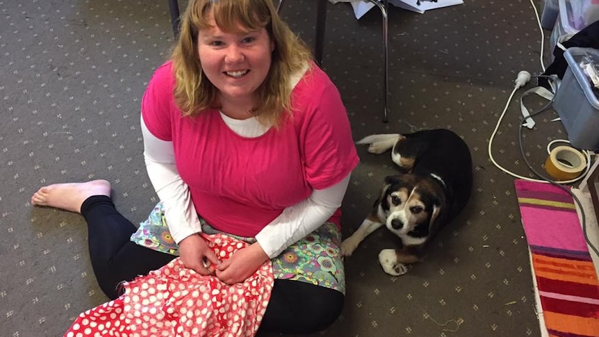 A young woman sitting on the ground, wearing pink, smiling with a small dog sitting next to her. 